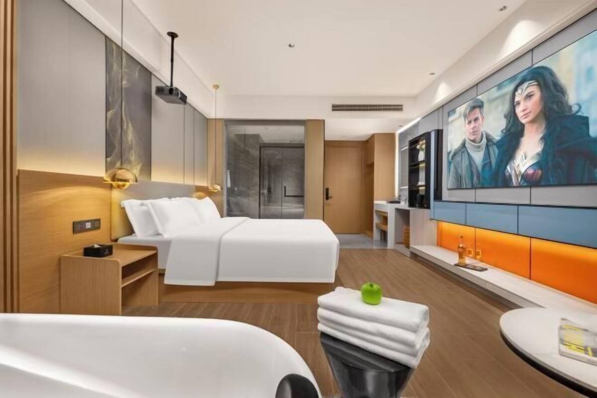 Une chambre d’hôtel moderne dotée d’un grand lit, d’un petit coin salon et d’un téléviseur fixé au mur qui diffuse une scène de film.