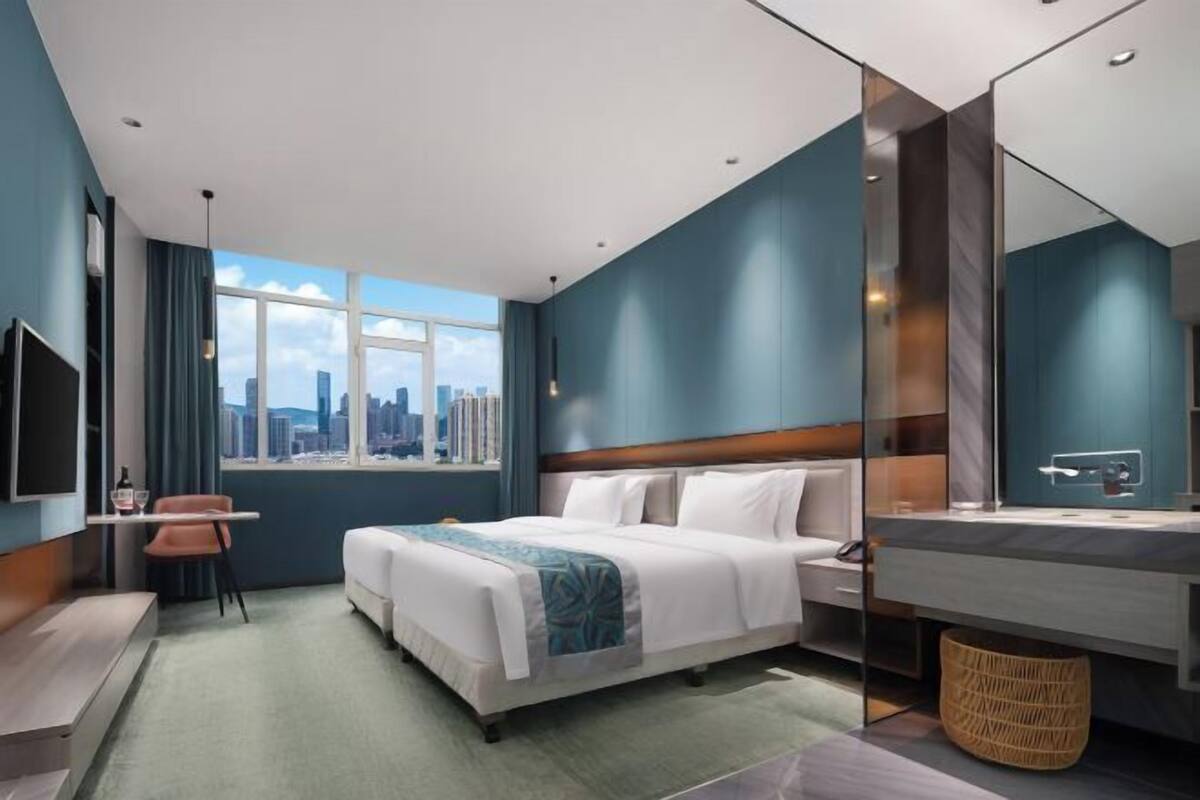 Une chambre d’hôtel moderne équipée d’un lit, d’un bureau, d’une chaise, d’une grande fenêtre donnant sur la ville, ainsi que d’une salle de bain avec un miroir et un lavabo.