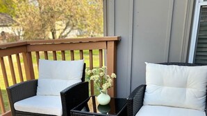 A deck with two wicker chairs and a small table, a vase with flowers, and a wooden railing.