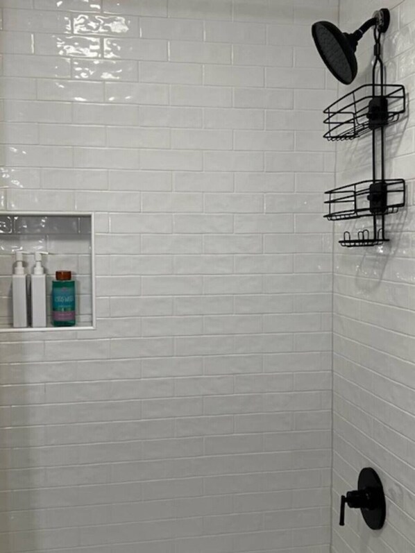 A bathroom with white tiled walls, a shower with a handheld showerhead, a wall-mounted faucet, and a shelf with toiletries.