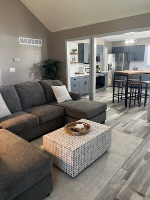 A modern living room with a sectional sofa, a patterned ottoman, and a kitchen in the background.