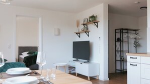 En moderne stue med et fladskærms-tv, et spisebord dækket til to personer og et køkkenområde med hvide skabe og en sort reol.