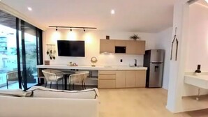 Une cuisine moderne équipée d’un téléviseur à écran plat, d’un coin repas avec une table et des chaises blanches, et d’une grande fenêtre offrant une vue sur la ville.