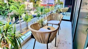 Un balcon avec des fauteuils en osier et une petite table, donnant sur une rue bordée d’immeubles et de palmiers.
