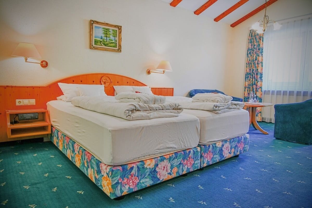 A hotel room with a double bed, a wooden headboard, a small bedside table with a lamp, a floral-patterned sofa, and a large window with curtains.