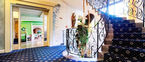 A grand staircase with ornate metal railings, a patterned carpet, and a statue of the Virgin Mary.