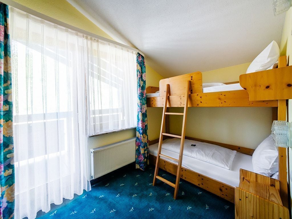 A small, single-bedded room with a wooden bunk bed, a ladder, a radiator, and a window with sheer curtains.