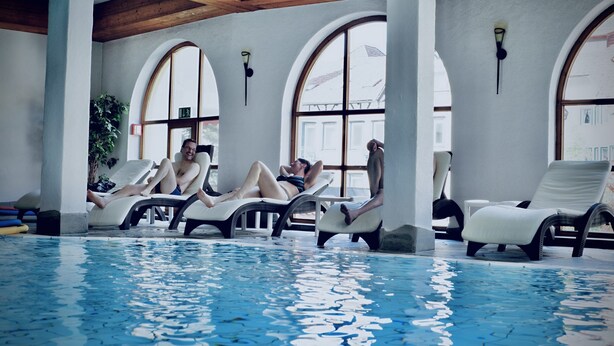 A pool area with lounge chairs and a view of the outside through large arched windows. - Holzschuh Schwarzwaldhotel (Baiersbronn)