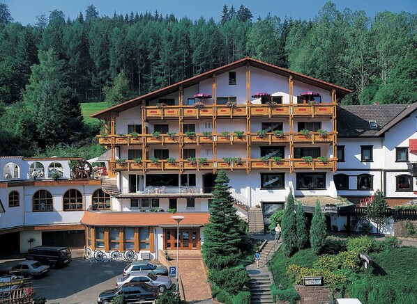 A multi-story hotel with balconies, a parking area, and a mountainous backdrop.