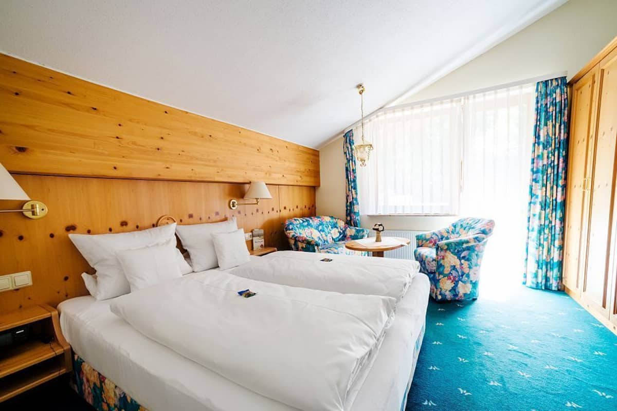 A hotel room with a wooden headboard, a bed with white linens, a small round table, a chair with floral upholstery, and a window with sheer curtains.