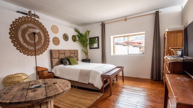 A bedroom with a bed, wooden floor, a round wooden table, a TV, and a window with a view of buildings outside. - Alojamiento en Centro Histórico Cusco (Cusco)