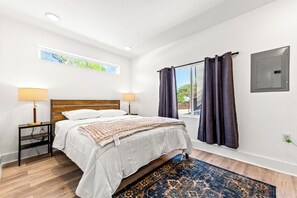 Une chambre à coucher avec un lit, deux lampes de chevet, une fenêtre avec des rideaux, une boîte électrique fixée au mur et un tapis à motifs.