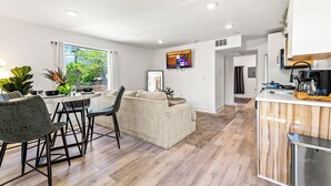 Una cocina moderna con encimera de madera, una zona de comedor con una mesa redonda y sillas, y una sala de estar con un sofá y un televisor montado en la pared.
