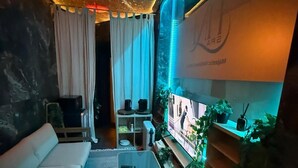 A modern living room with a sofa, coffee table, and TV. There are plants and a shelf with decorative items.