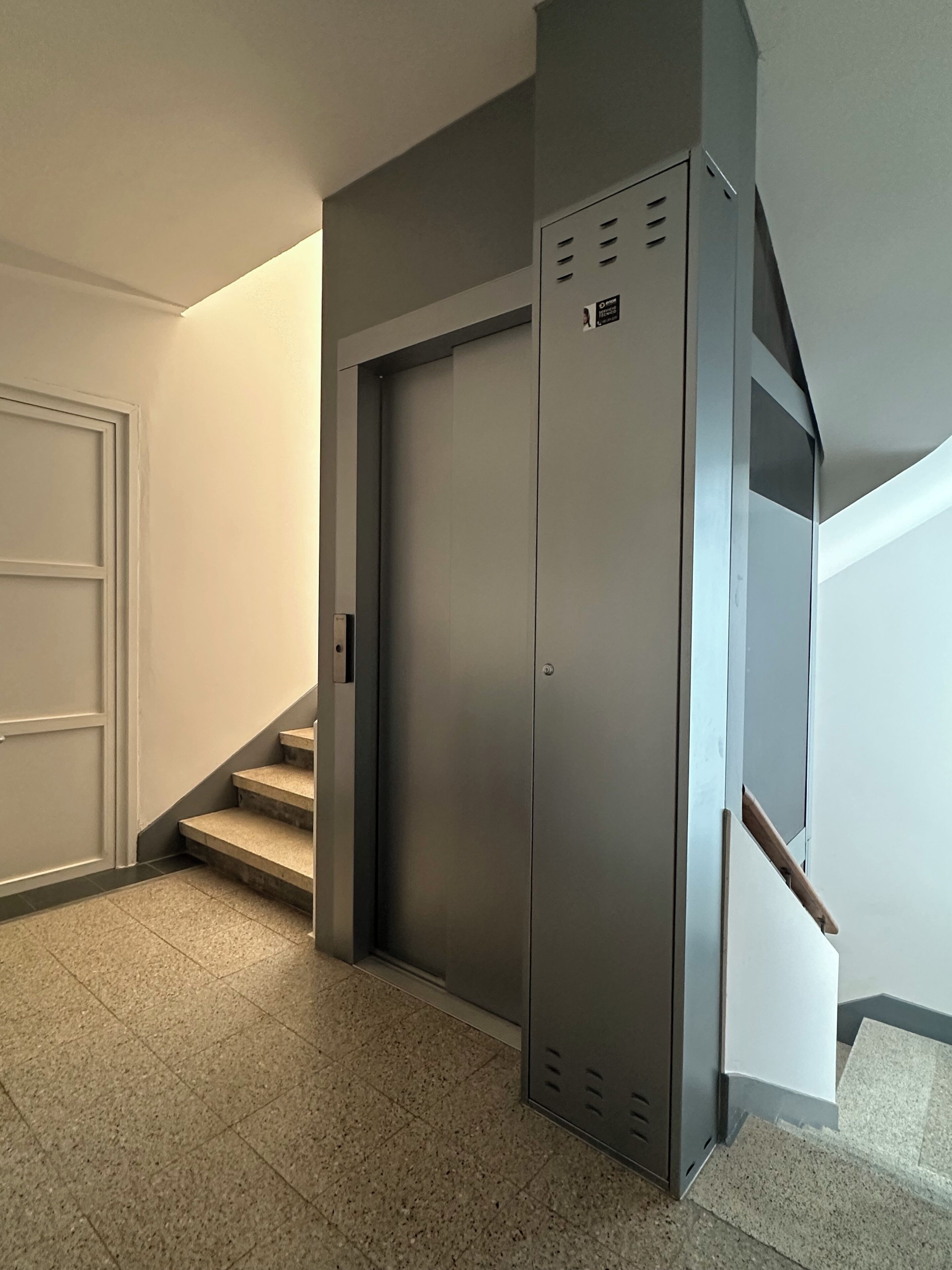 A modern elevator with a metallic finish, located in a hallway with a door and a staircase.