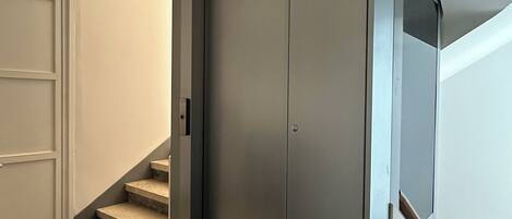 A modern elevator with a metallic finish, located in a hallway with a door and a staircase.
