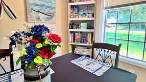 Un comedor con una mesa dispuesta para dos, un florero con flores, una estantería con diversos objetos y una ventana con vistas a la vegetación.