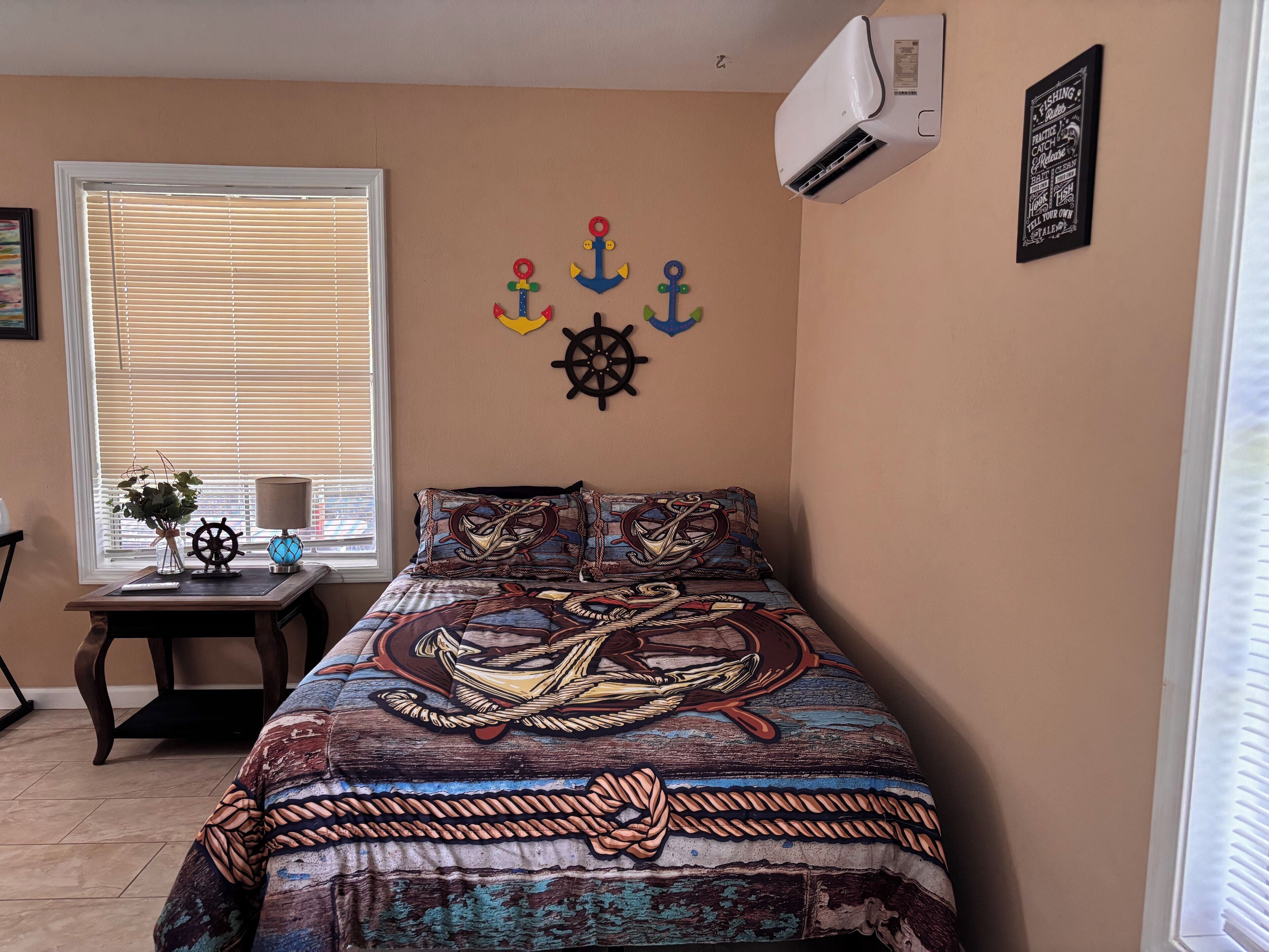 A bedroom with a bed featuring a nautical-themed bedspread, a window with blinds, a small table with a lamp and a vase of flowers, and wall decorations including anchors and a ship's wheel.
