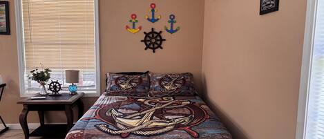 A bedroom with a bed featuring a nautical-themed bedspread, a window with blinds, a small table with a lamp and a vase of flowers, and wall decorations including anchors and a ship's wheel.