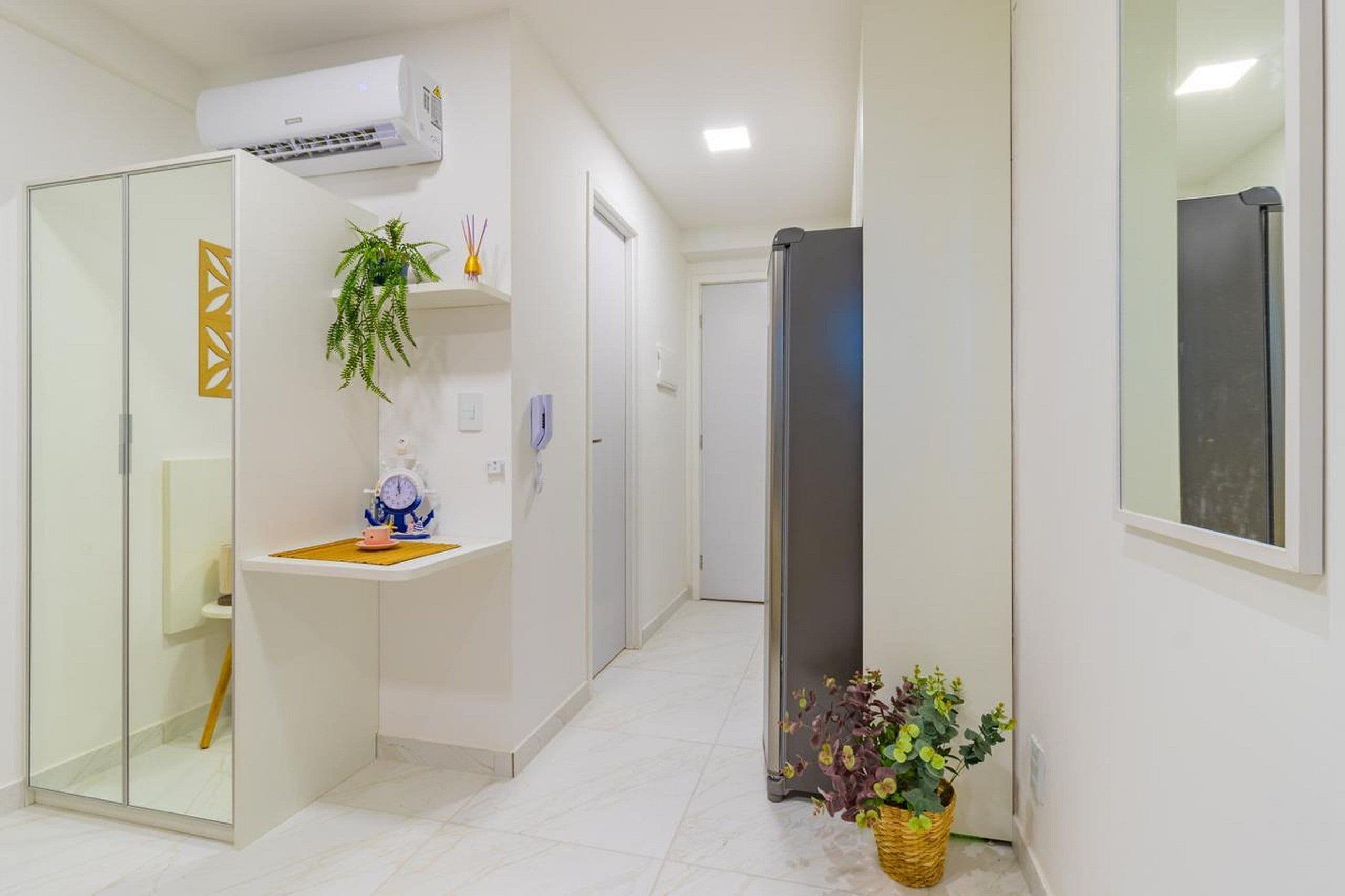 Un pasillo con suelo de baldosas blancas, una puerta de vidrio, una pared blanca con un panel decorativo, una planta en maceta y un equipo de aire acondicionado instalado en la pared.