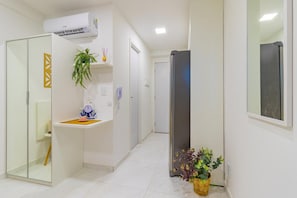 A hallway with a white tiled floor, a glass door, a white wall with a decorative panel, a potted plant, and a wall-mounted air conditioning unit. - Cupe Garden - Estúdio Superior (Ipojuca)