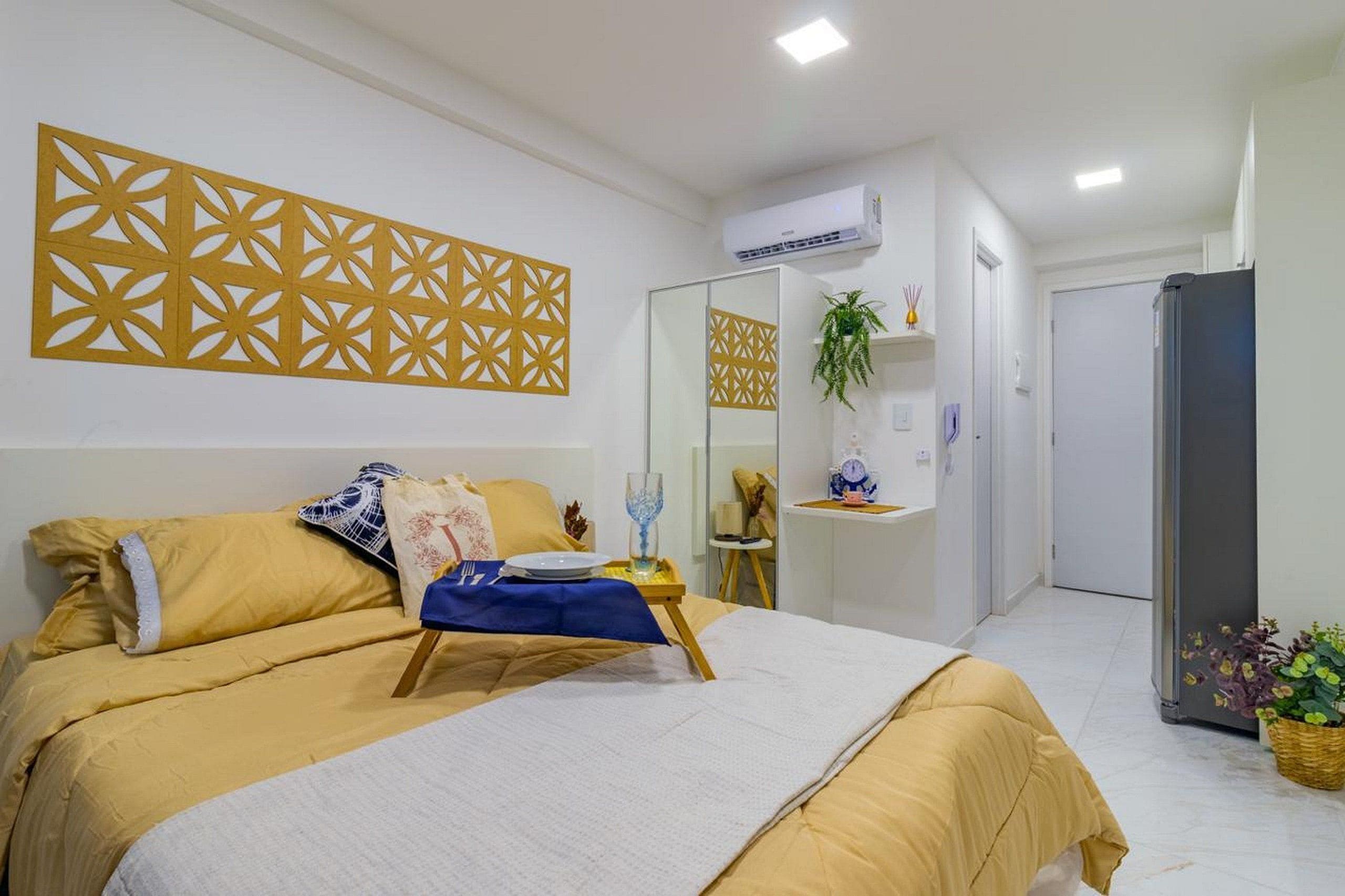 Quarto de hotel com uma cama, uma mesa de cabeceira com tecido azul, um painel decorativo na parede, um aparelho de ar condicionado, um espelho, uma planta e um frigorífico.