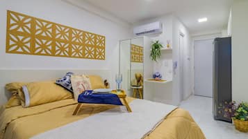 Quarto de hotel com uma cama, uma mesa de cabeceira com tecido azul, um painel decorativo na parede, um aparelho de ar condicionado, um espelho, uma planta e um frigorífico.