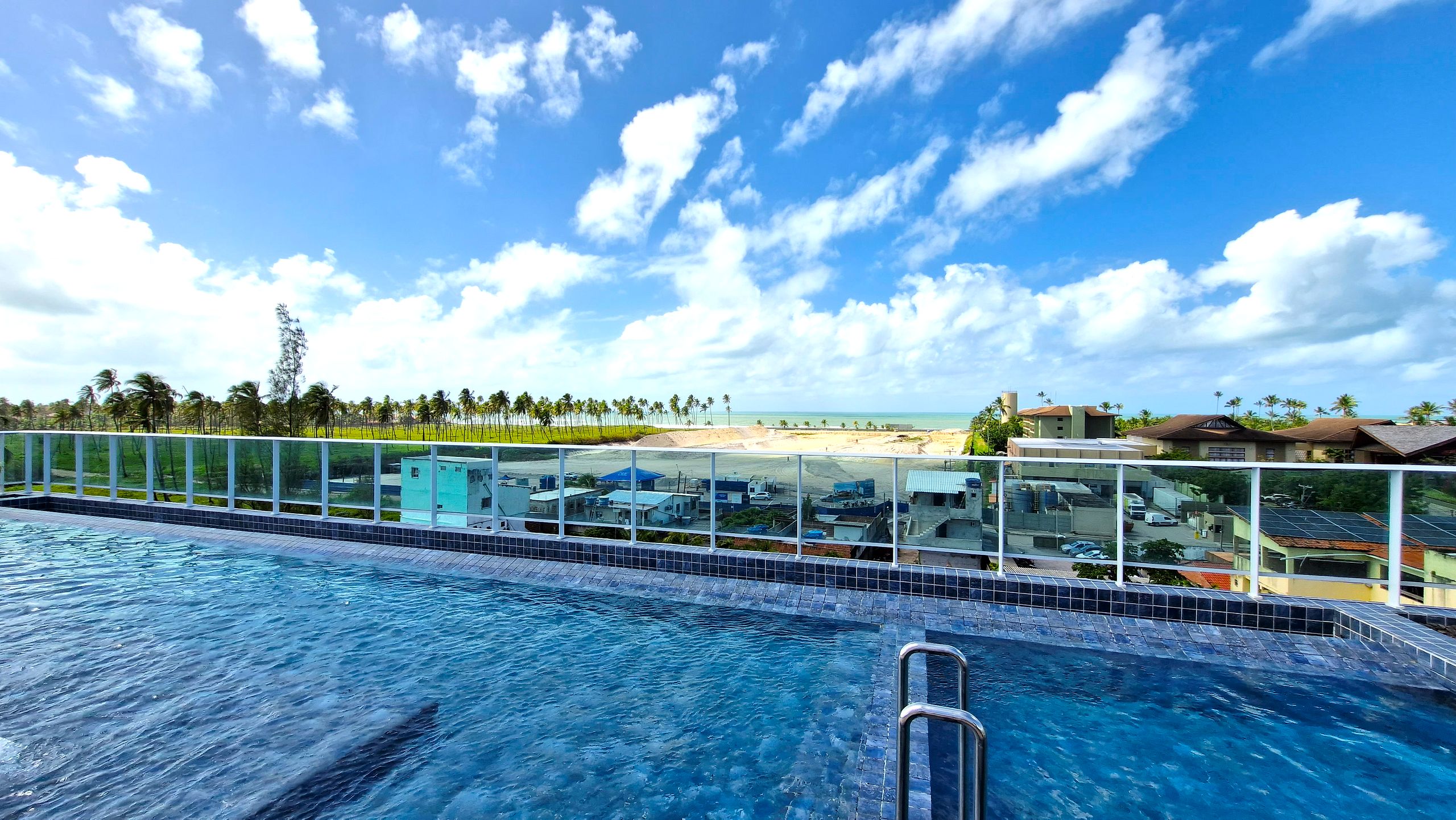 Piscina com guarda-vidro, vista para a praia e palmeiras.