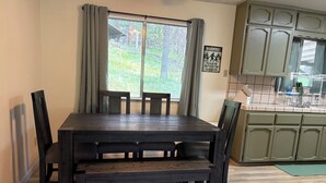 A dining area with a dark wooden table and chairs, a window with curtains, and a kitchen area with green cabinets and a sink. - Bigfoot Lodge Yosemite Bass Lake (Oakhurst)