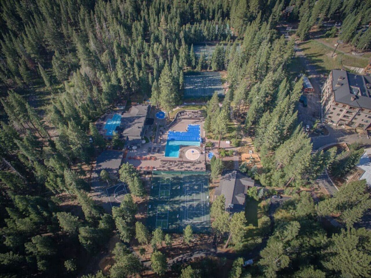 An aerial view of a property surrounded by dense forest, featuring a swimming pool, tennis courts, and a small building.