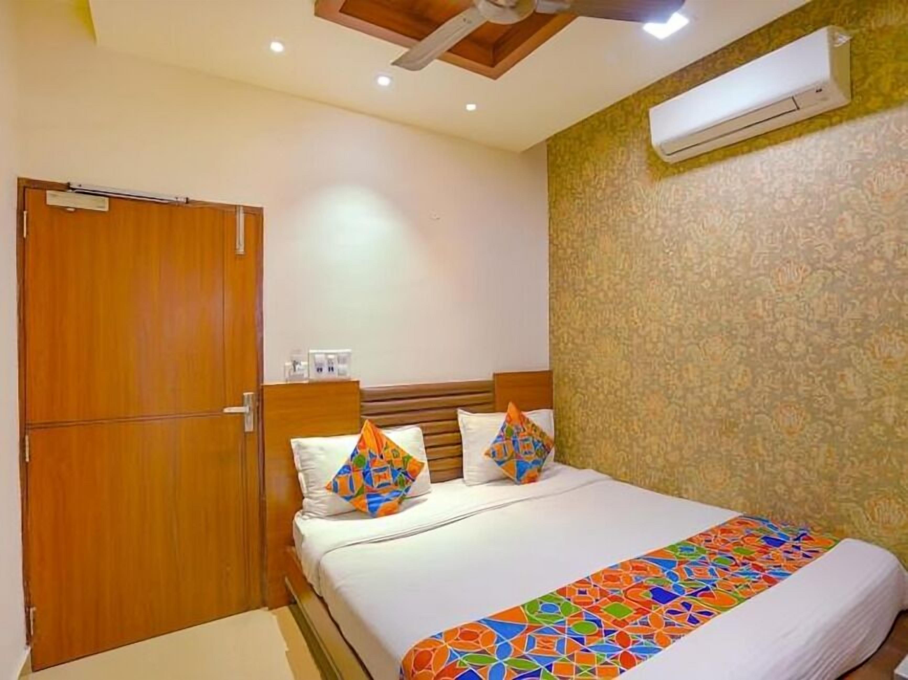 A hotel room with a wooden door, a bed with colorful pillows, a patterned wall, and an air conditioning unit.
