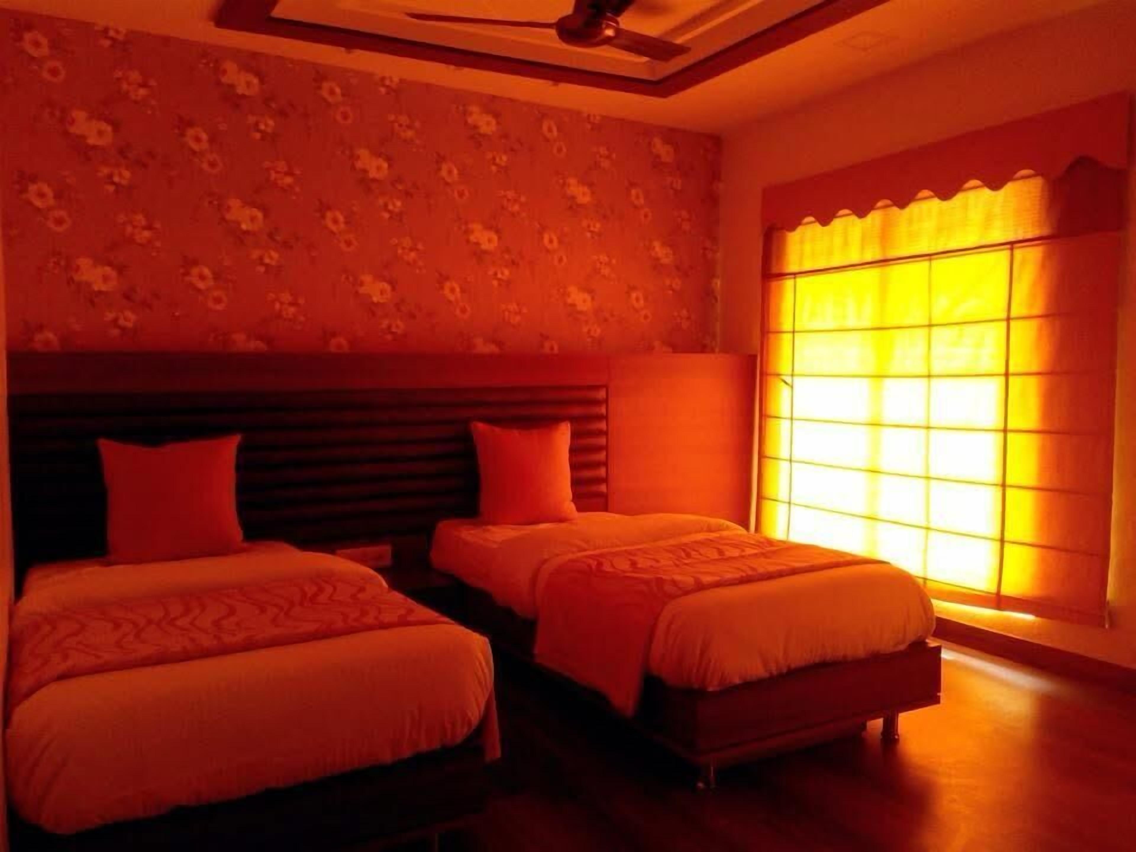 Habitación de hotel con dos camas, ventilador de techo, papel tapiz floral y un amplio ventanal con cortinas.