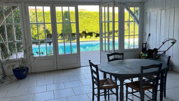 Un patio techado con una mesa y sillas, una planta en maceta y vistas a una piscina y follaje.