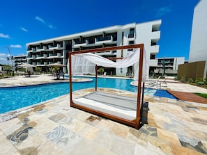 A poolside area with a wooden canopy bed, a swimming pool, and a modern building in the background. - Makia Beach Experience - A117 (Ipojuca)