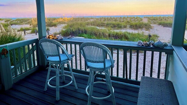 A covered outdoor area with two wicker chairs and a small table, overlooking a beach with sand dunes and a clear sky.