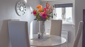 Un comedor con una mesa redonda, sillas, un jarrón de flores, un reloj de pared y una ventana con vistas a un castillo.