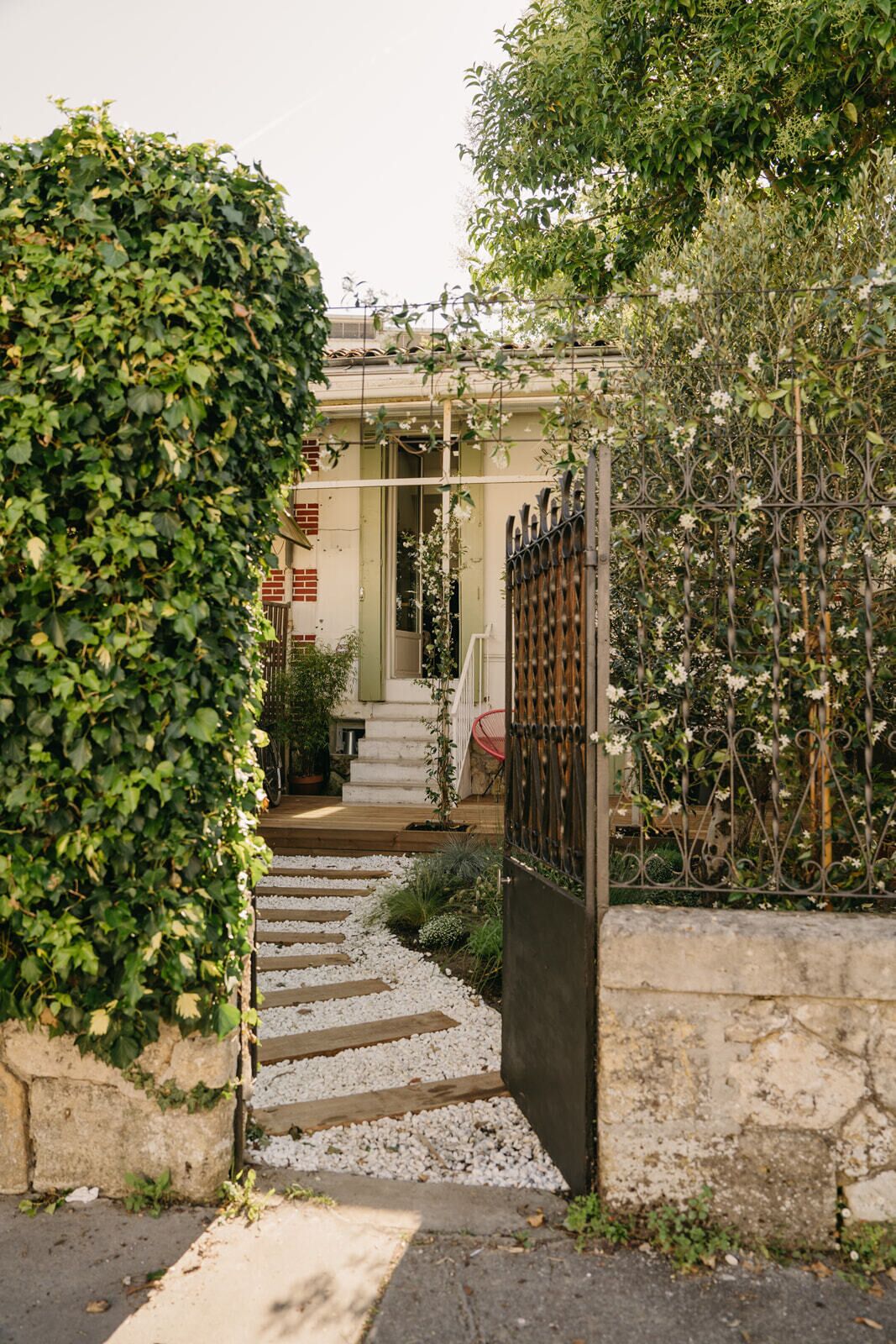 A garden entrance with a stone wall, a metal gate, and a pathway leading to a building with a green door.