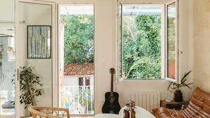 A living room with a wicker chair, a brown leather sofa, a small table, and a guitar. There are two windows with a view of greenery outside. - Charming renovated house with small pool in the La Bastide district (Bordeaux)