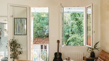 A living room with a wicker chair, a brown leather sofa, a small table, and a guitar. There are two windows with a view of greenery outside.