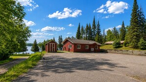 A gravel driveway leading to a red building with a small shed, surrounded by trees and a lake.