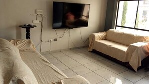 A room with a flat-screen TV mounted on the wall, a small table with cups, a sofa, and a window providing natural light.