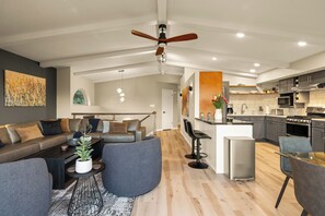 A modern living room with a sofa, coffee table, and chairs, a kitchen with appliances, and a ceiling fan.