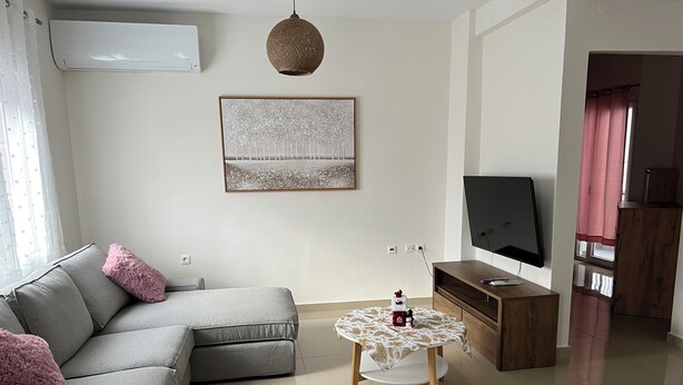 Ein modernes Wohnzimmer mit grauem Sofa, einem kleinen runden Tisch mit einem Weinglas, einem Flachbildfernseher auf einem Holzboden und einer Wanddekoration.