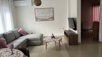 Een woonkamer met een grijze bank, een klein rond tafeltje met een witte tafelkleed, een televisie op een houten dressoir, en een schilderij aan de muur.