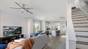 En rummelig opholdsstue med sofa, TV og loftventilator. En trappe fører til en etage ovenpå.