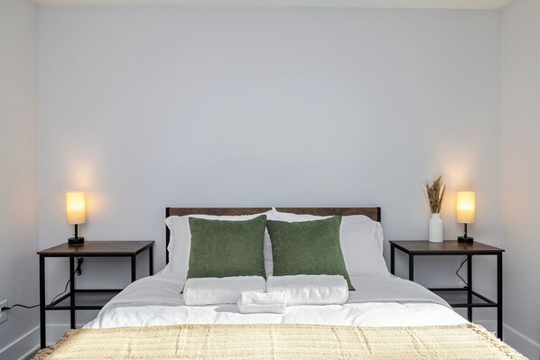Una cama bien hecha con ropa de cama blanca, dos almohadas verdes y una manta color beige. Hay dos mesitas de noche, cada una con una lámpara y un jarrón con plantas secas.