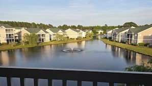 A lakeside view of a residential area with multiple two-story buildings, a central fountain, and a clear sky. - Sandpiper Bay 3rd Floor. 4 mi to beach. 3 bd, 2 ba, new kitchen,  pool & more (Sunset Beach)
