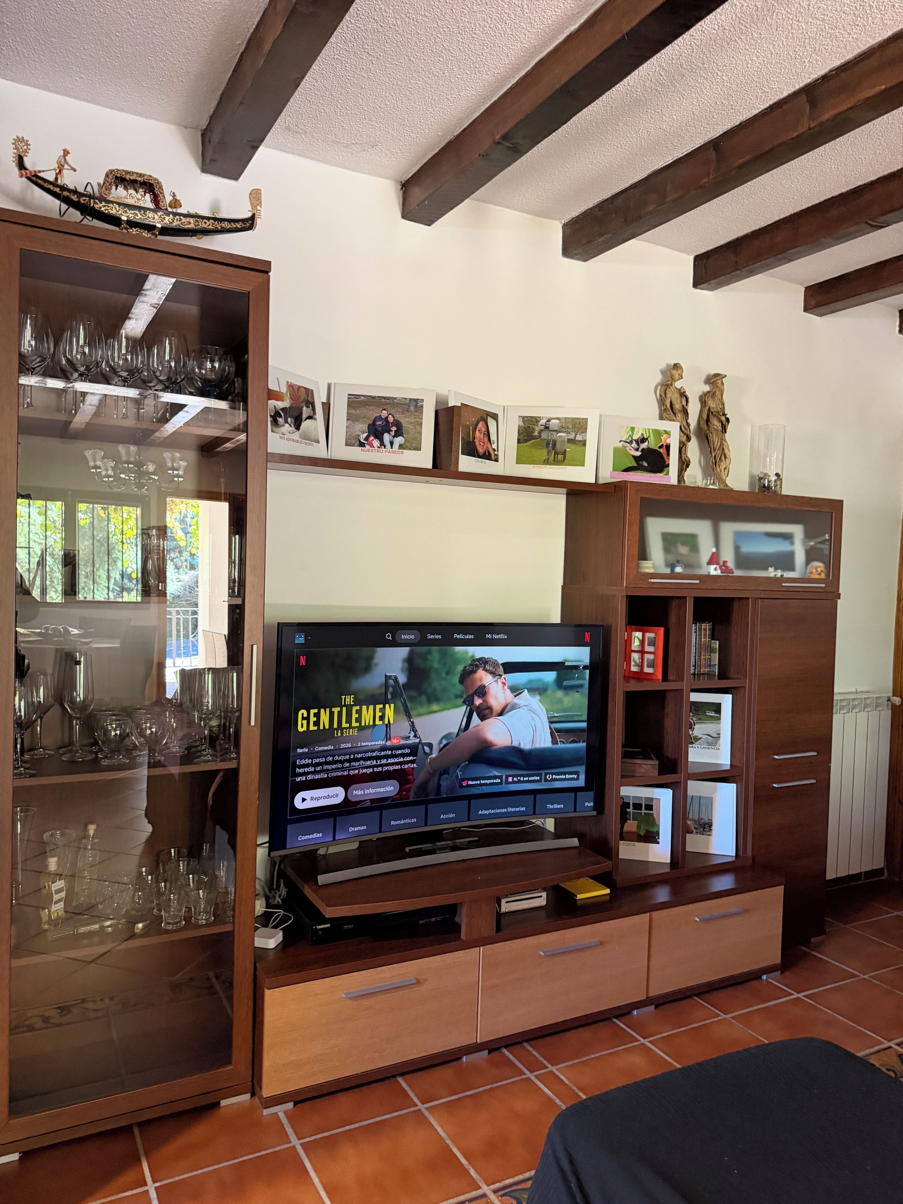 A living room with a flat-screen TV mounted on a wooden entertainment unit, a glass cabinet with wine glasses, and a ceiling with exposed beams.
