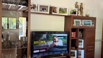 A living room with a flat-screen TV mounted on a wooden entertainment unit, a glass cabinet with wine glasses, and a ceiling with exposed beams.