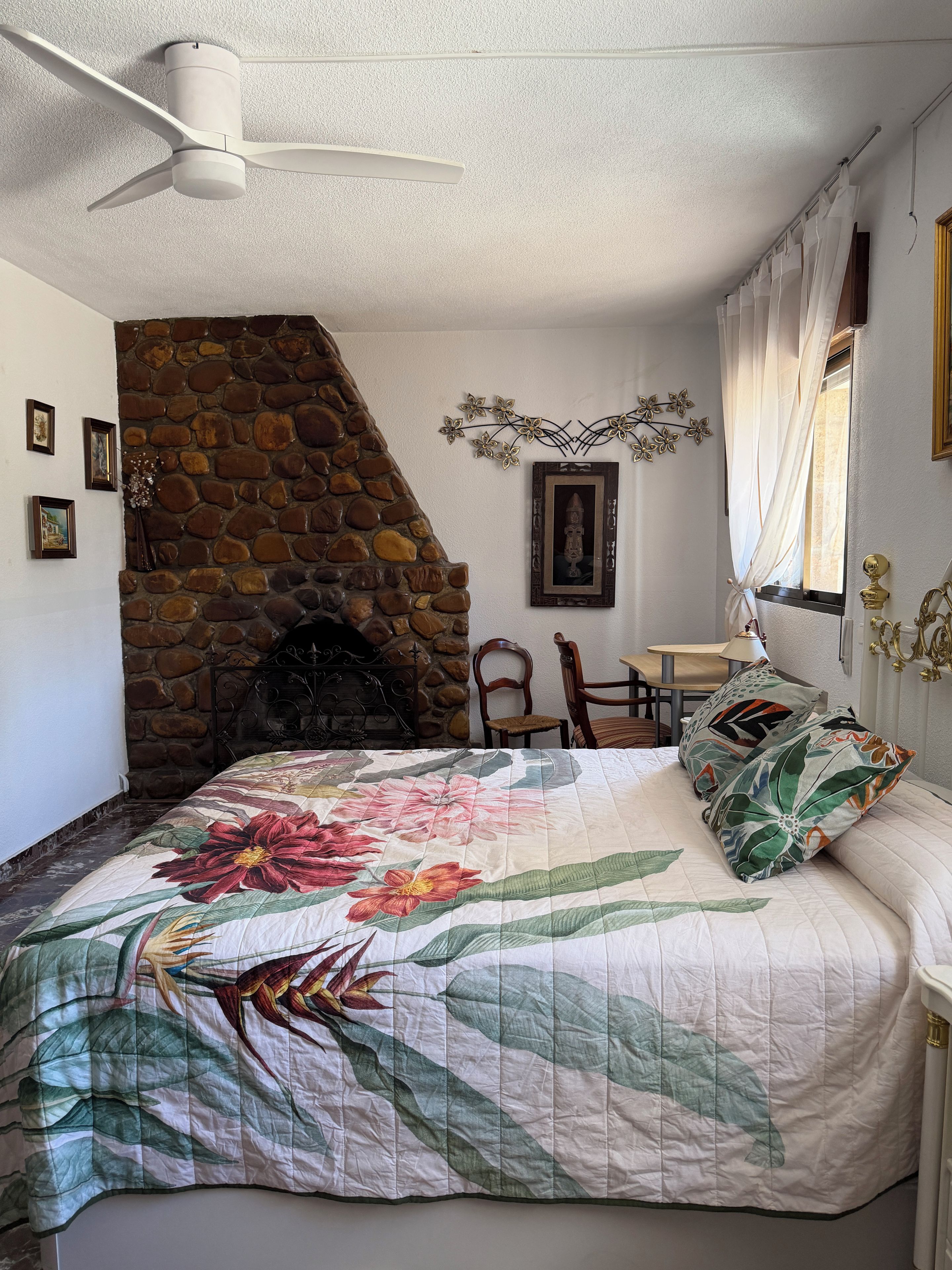 Ein Schlafzimmer mit Kamin aus Stein, einem Bett mit floralem Bettbezug, einem Deckenventilator und einer Essecke mit Tisch und Stühlen.